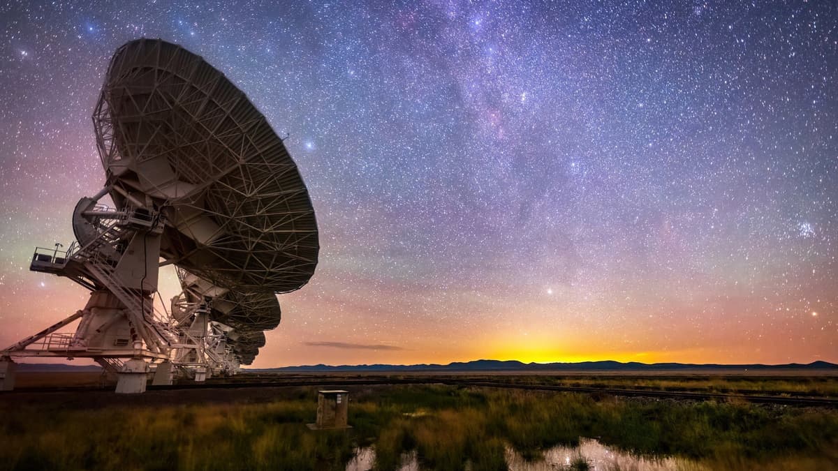 Night view of the Very Large Array radio telescope in New Mexico: antennas on the left against the Milky Way, a sunset glow on the horizon, a puddle reflecting the stars in the grass, and the Pleiades star cluster at right.