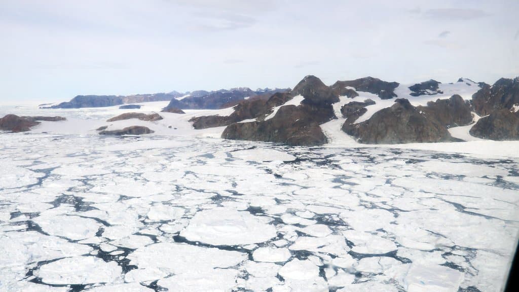 Aerial view of Antarctic sea ice broken into floes, with dark open water in the cracks between them and a rocky, snow-covered coast behind.