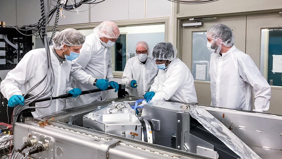 Five engineers in white clean-room suits and face masks lean over a long metal vacuum chamber holding the NEID spectrograph.