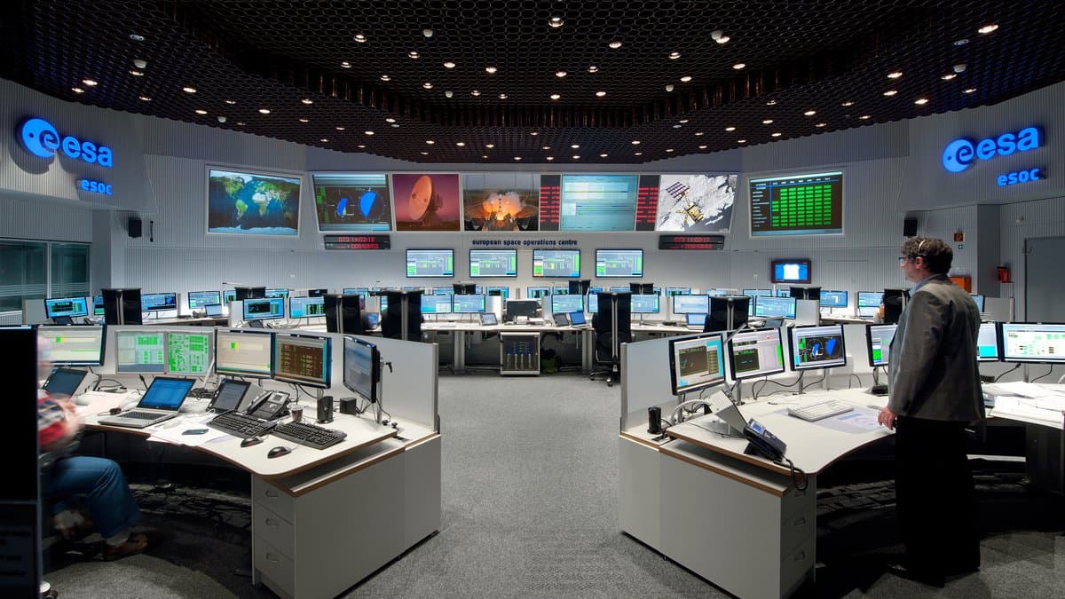 Operators at banks of consoles in a large spacecraft operations center, with world maps and telemetry displays on the far wall.