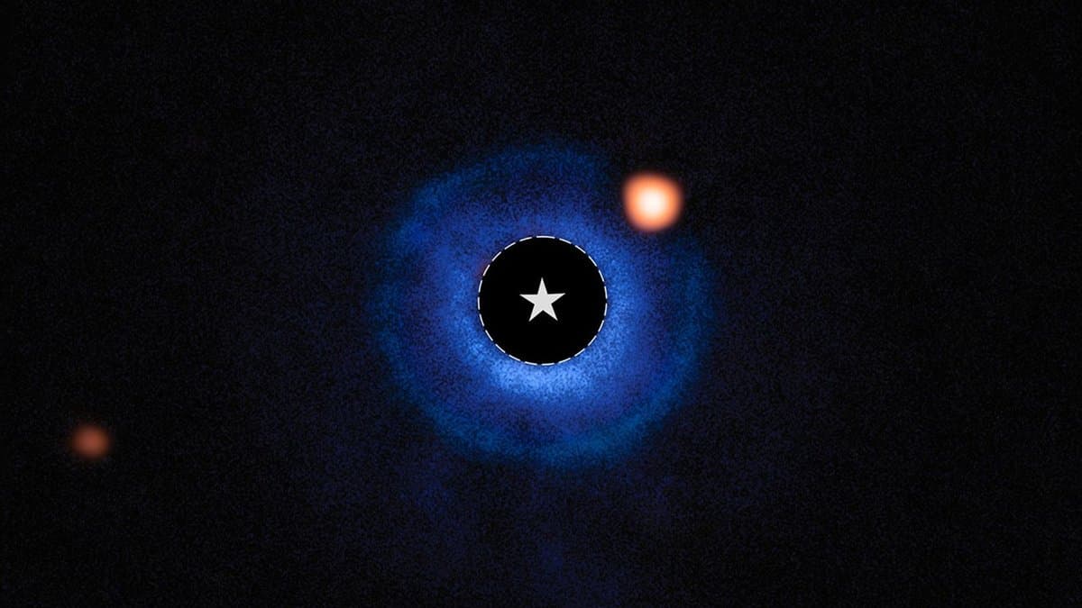 A coronagraph image in which the central star is hidden behind a dark mask marked with a star symbol, ringed by blue dust, with an orange point of light just outside the ring at upper right.