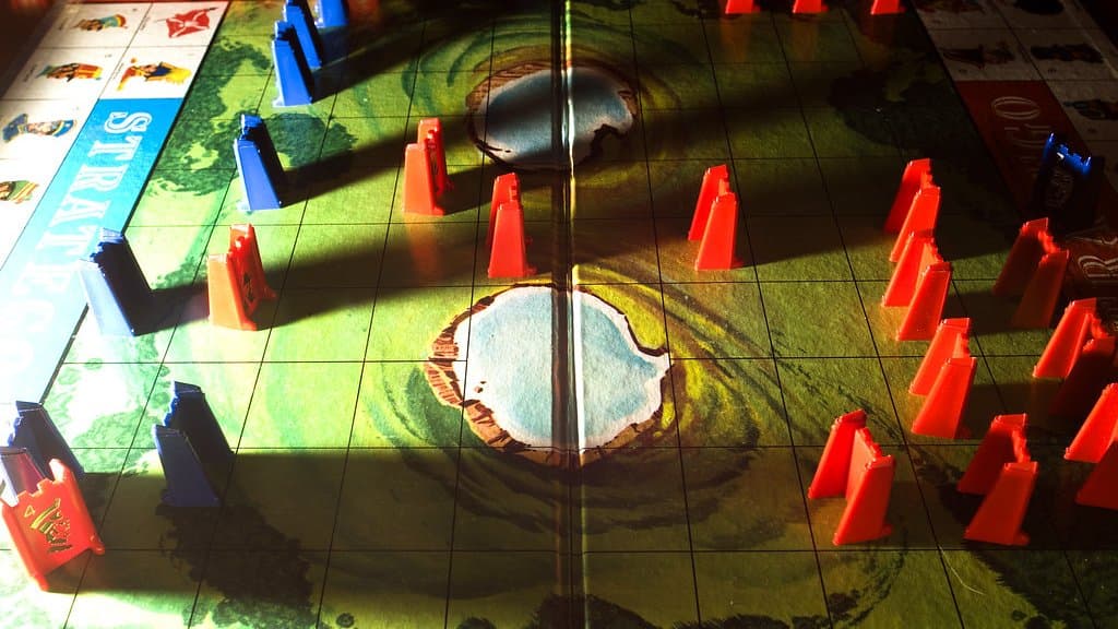 Red and blue plastic Stratego pieces standing on a game board, with two painted lakes in the middle of the battlefield.
