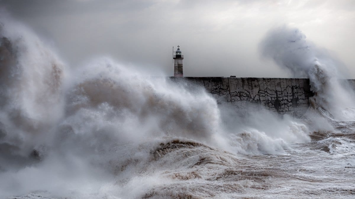 A massive storm-driven wave breaking over a lighthouse and harbour wall on the UK coast