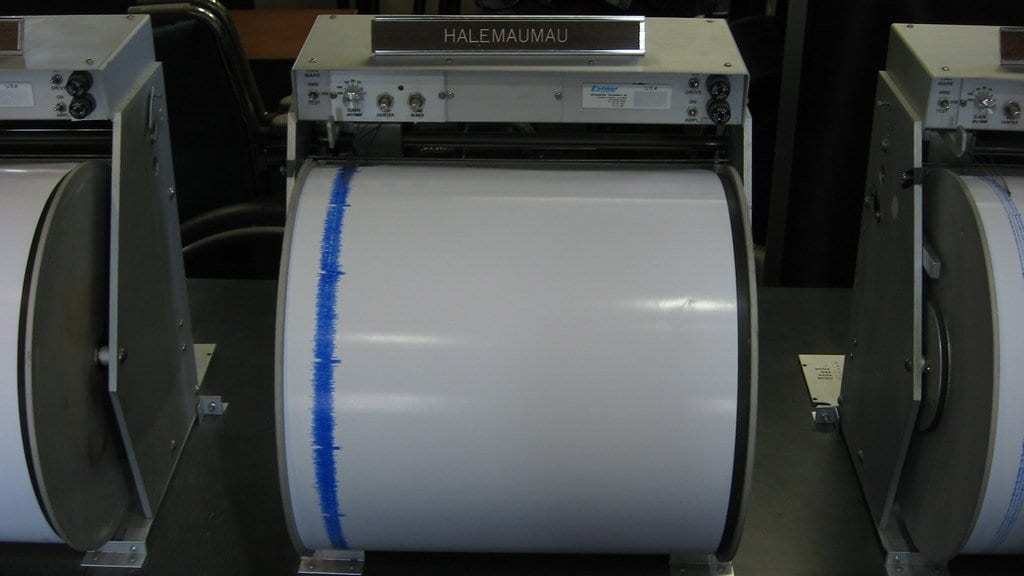 Three analogue paper-drum seismograph recorders in a row on a dark bench in a museum display; the central recorder carries an engraved HALEMAUMAU nameplate and its white paper drum shows a single ragged blue ink trace