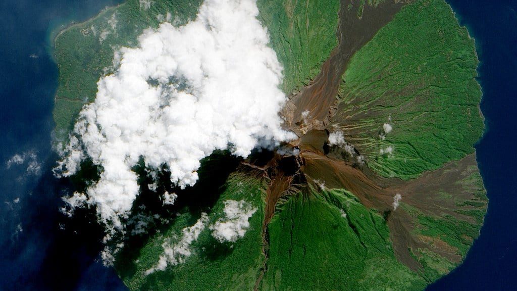 A satellite view of an erupting island volcano venting a plume over the surrounding ocean.
