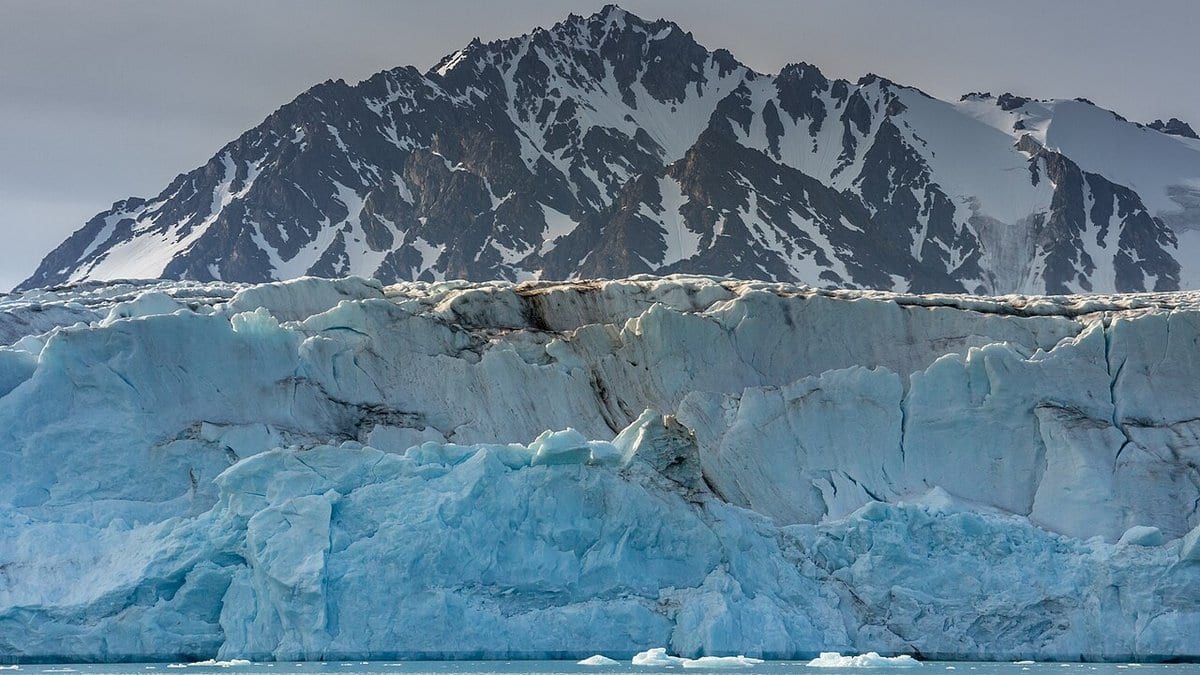 The blue ice cliff of a tidewater glacier meeting the sea in Svalbard, with a dark snow-streaked mountain rising behind it.