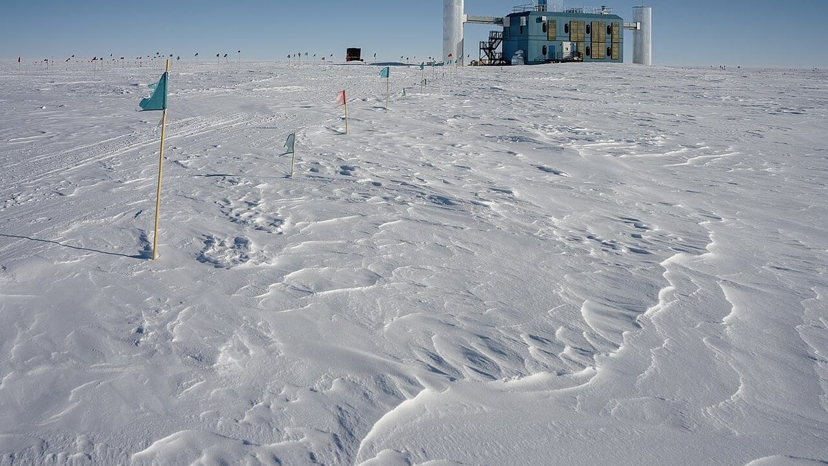 The IceCube Laboratory, a boxy building raised on stilts, standing alone on the flat Antarctic ice sheet at the South Pole, with survey flags in the foreground.