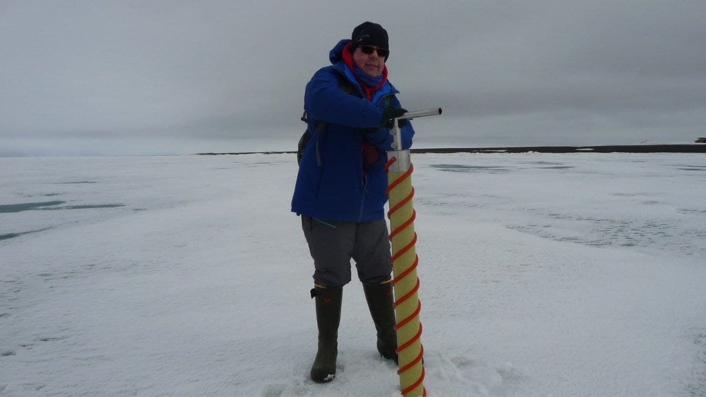 A researcher in a blue parka stands on flat sea ice holding a long spiral ice corer, with open water and a low shoreline behind