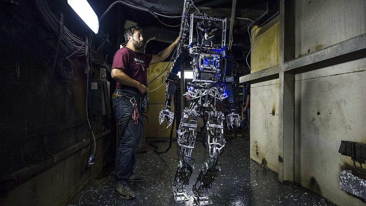 A researcher steadies a life-size bipedal humanoid robot by its frame during a trial in a narrow steel corridor