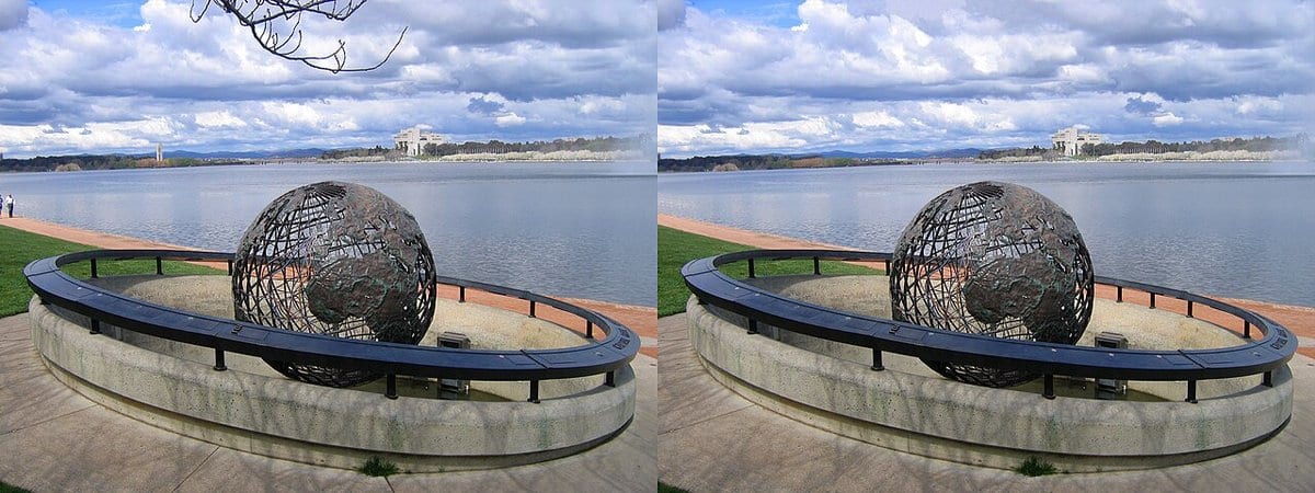Two near-identical photographs placed side by side: a large open lattice globe sculpture on a curved stone plinth beside a lake, with a low pale public building and low hills on the far shore under a cloudy sky. Small details differ between the frames, including a bare branch that hangs into the sky at the top left of the left-hand image and is absent from the right-hand one.