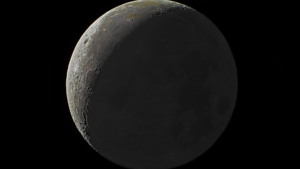 Telescopic photograph of the Moon: a narrow sunlit crescent along the left limb, with the rest of the near-side disc showing faintly in earthshine against a black sky.