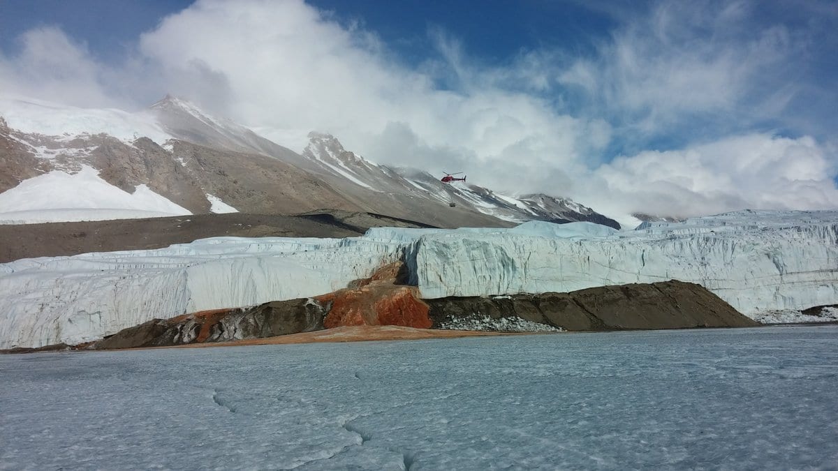 The snout of Taylor Glacier in Antarctica: a white ice cliff meeting bare rock, with a rust-red stain spilling down the ice and over the moraine below it. A small red helicopter hovers against snow-covered mountains and cloud behind, and the frozen surface of the lake fills the foreground.