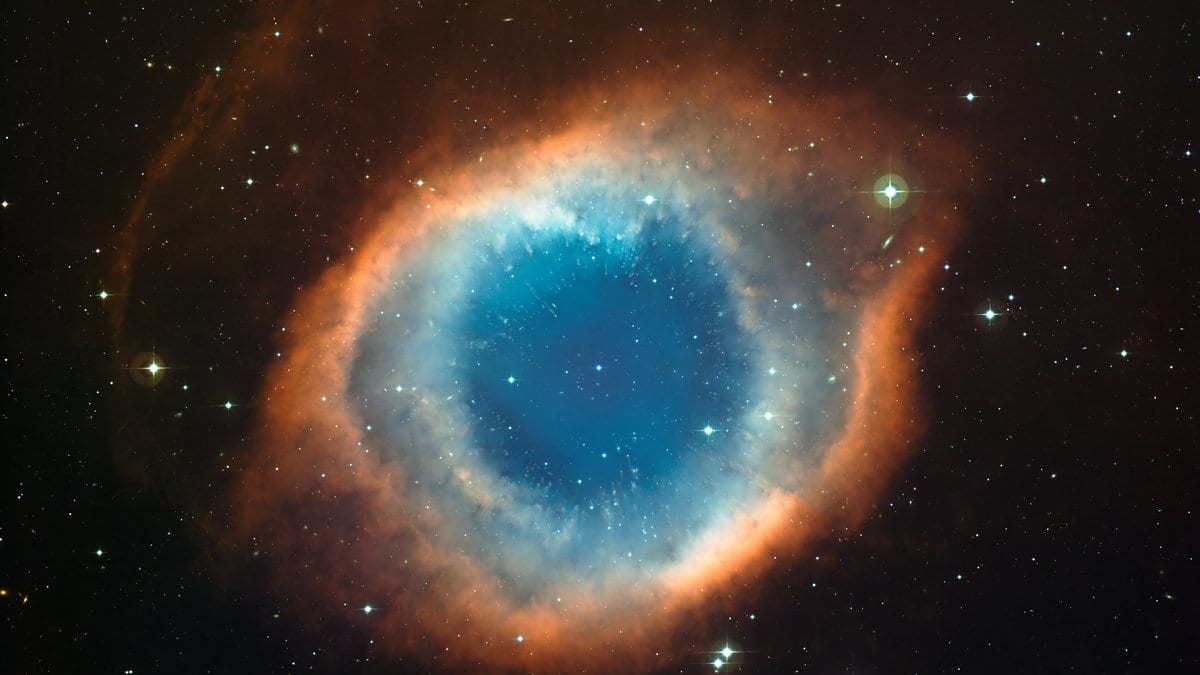 Wide-field telescope image of the Helix Nebula: a bright blue-green ring of glowing gas with a red outer rim, set against a dense star field.