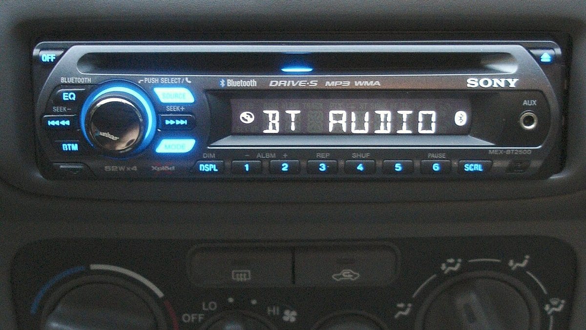 An illuminated aftermarket head unit fitted into a car dashboard, its display reading BT AUDIO above a row of preset buttons and heater controls.