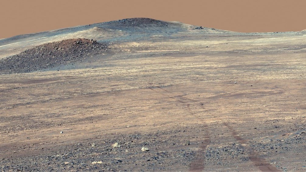 Wide view across a pale, dust-covered Martian slope, with a broad band of bright, light-toned rock running from the left of the frame to the right beneath darker, rubbly ridges, and faint rover wheel tracks curving down the slope in the foreground.