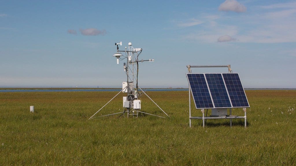 A solar-powered instrument mast standing on flat coastal tundra beside a bank of solar panels, under a pale Arctic sky
