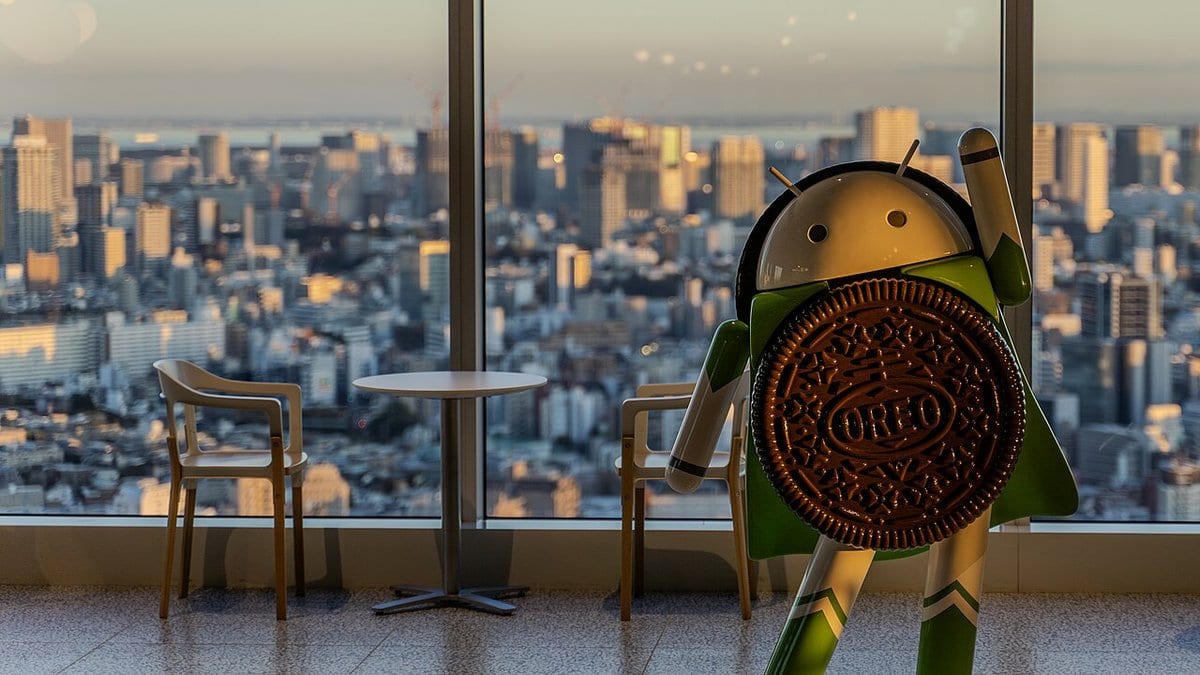A statue of the Android robot mascot holding an Oreo biscuit, standing by a window above a city skyline at dusk