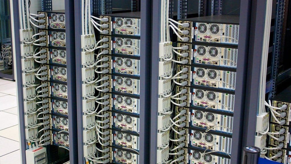 Rows of rack-mounted server units stacked floor to ceiling in a data centre aisle, each unit trailing pale network and power cables.