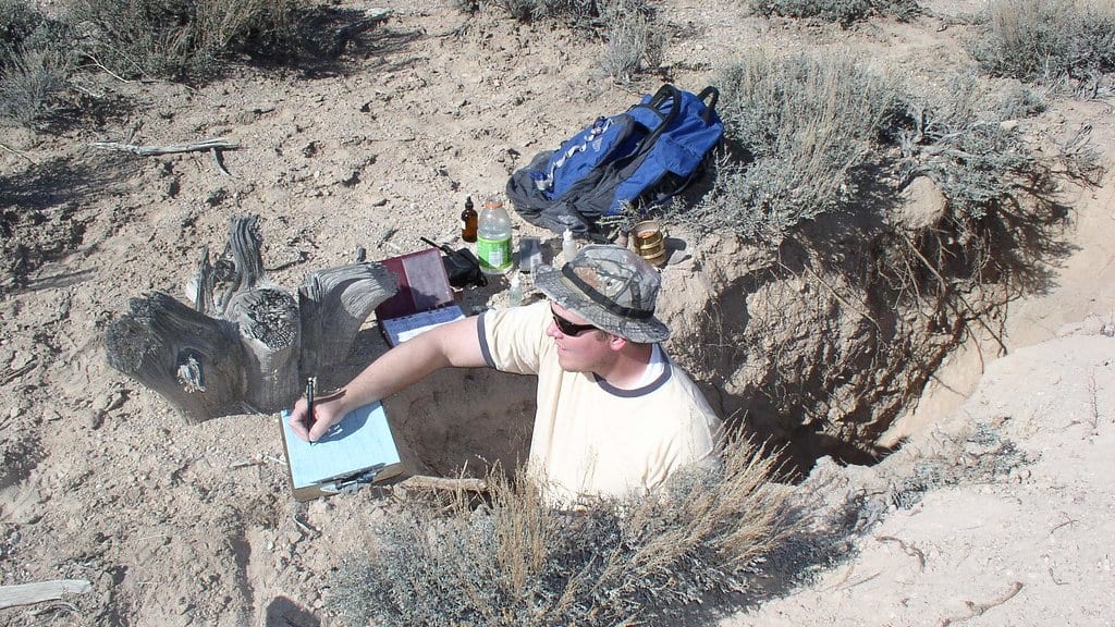 A soil scientist standing waist-deep in a freshly dug soil pit, writing notes on a clipboard beside sampling gear.