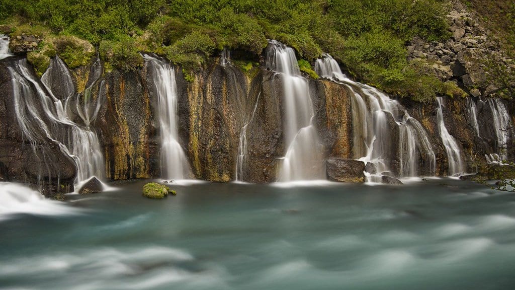 A long line of small waterfalls emerging from beneath a mossy lava field and spilling into a wide turquoise river.