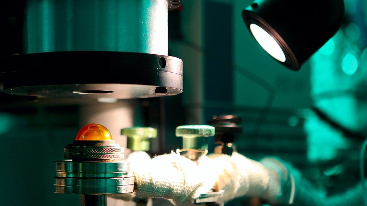 Close-up of the sample stage of a diffuse-reflectance infrared spectrometer, an amber sample dome and knurled adjusters lit under a lamp in a laboratory.
