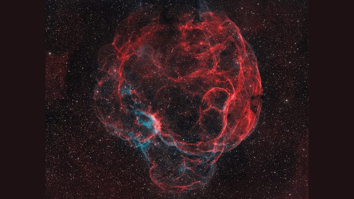 A large, faint supernova remnant filling the frame: a rough sphere of tangled red hydrogen filaments laced with blue-green oxygen emission, set against a dense field of stars.