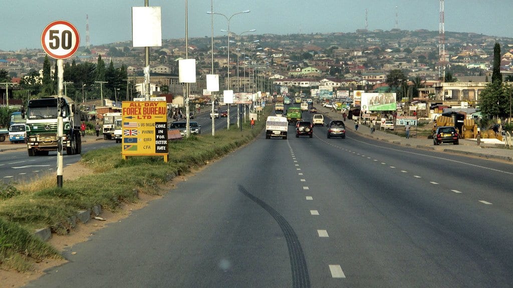 A multi-lane road on the edge of Accra carrying trucks, minibuses and cars, with roadside kiosks and advertising boards on the left and densely packed low-rise houses covering the hillside behind.