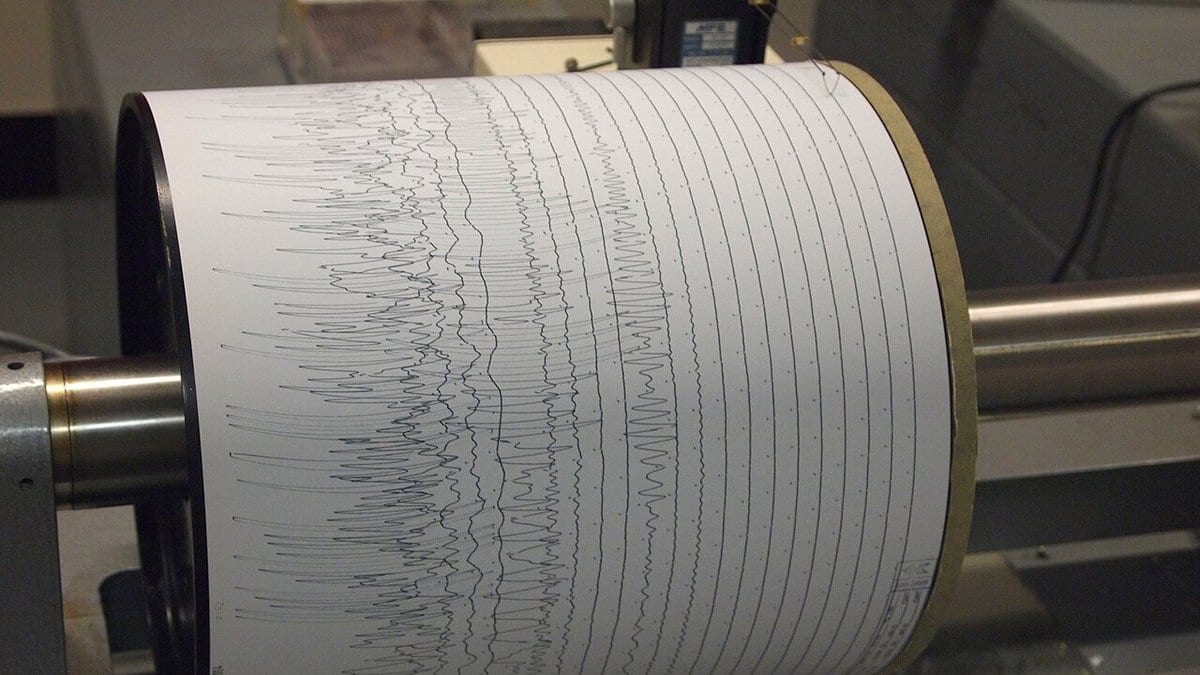 Close-up of a rotating drum seismograph with paper wrapped around it, covered in rows of inked seismic traces including dense bursts of rapid oscillation.