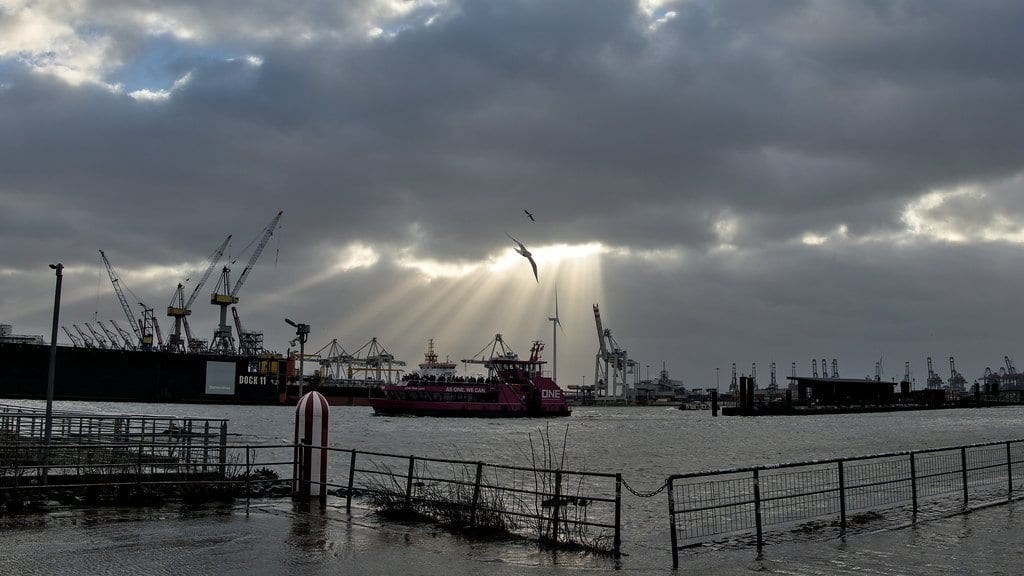 Floodwater covers a fenced riverside promenade in the Port of Hamburg during a storm surge, with container cranes, a ferry and sunbeams breaking through heavy cloud behind.