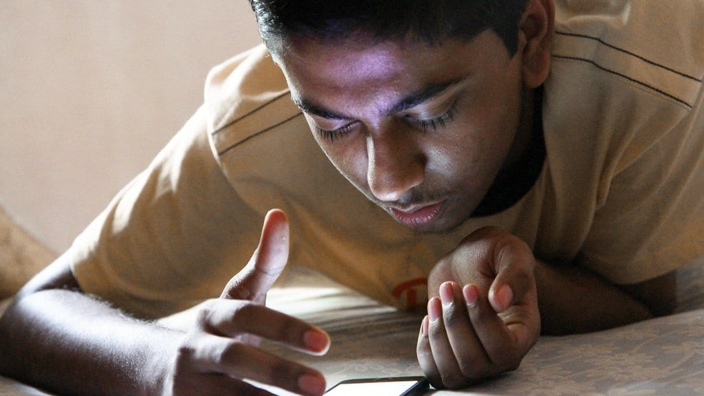 A teenage boy lies on a bed reading a smartphone at close range, his face lit by the screen.