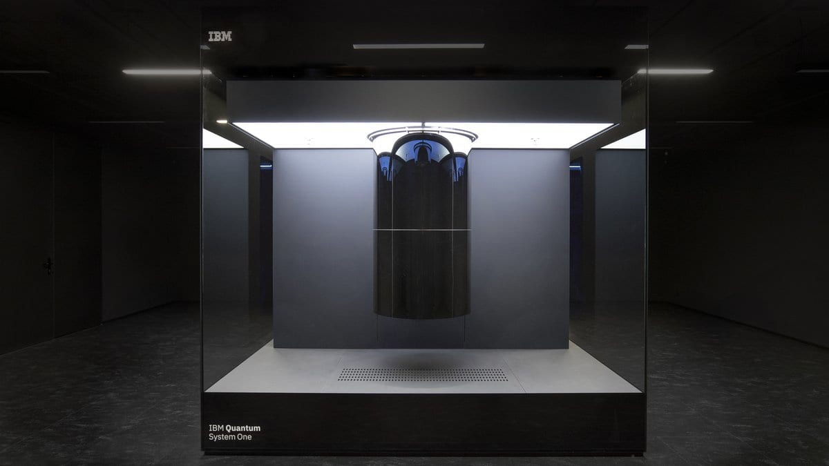 An IBM Quantum System One: a cylindrical cryostat suspended inside a sealed glass cube in a darkened room, with the IBM Quantum System One nameplate on the plinth.