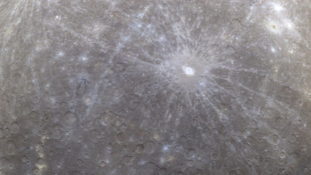Mercury's gray, heavily cratered surface fills the frame in an orbital view, with a bright rayed impact crater and long, pale rays stretching across older cratered plains.