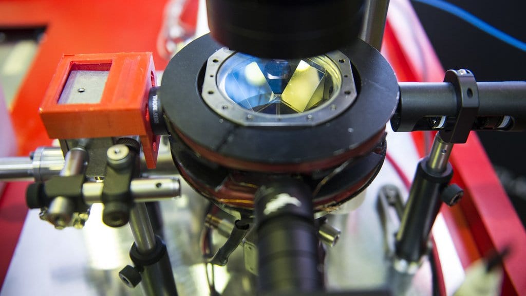 Close-up of an atom interferometer: a stainless-steel ultra-high-vacuum chamber with a bolted viewport window, surrounded by laser optics on adjustable mounts.
