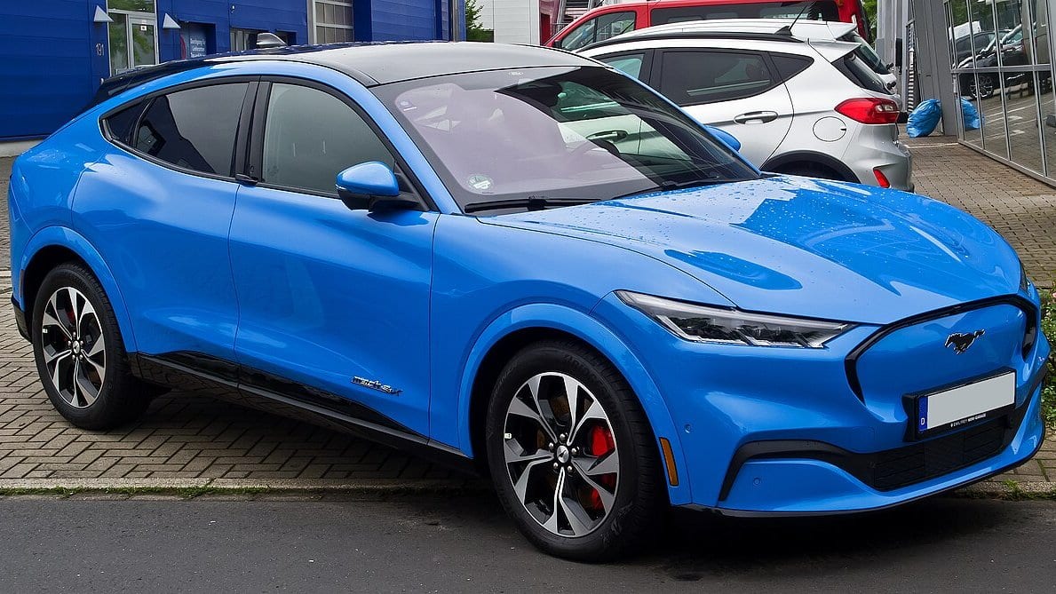 A blue Ford Mustang Mach-E all-electric SUV parked at a kerb, photographed from the front three-quarter angle.