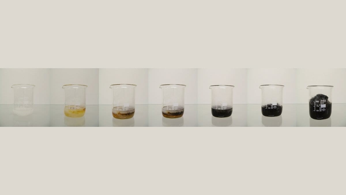Seven glass beakers photographed in sequence: clear liquid on the left darkens through yellow and brown to a solid black column of carbon on the right.