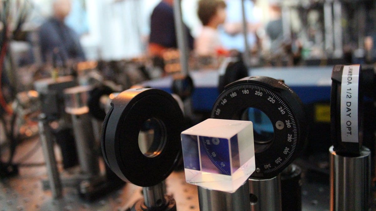 Optical elements on a laser table: a transparent beamsplitter cube in front of a black lens mount and a graduated rotation stage, with more mounted optics receding into the background.