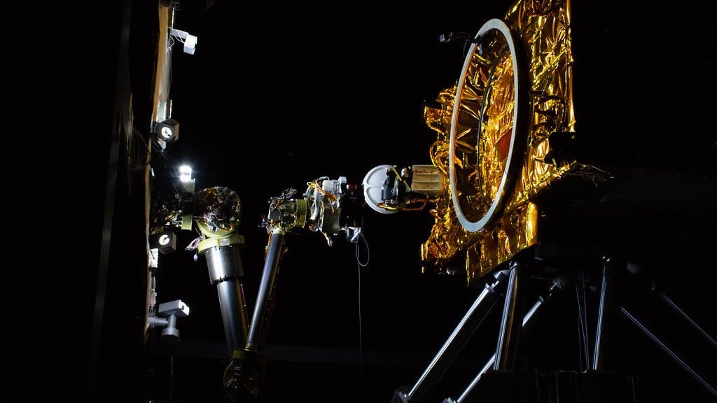 A robotic arm extends toward a gold-foil-wrapped satellite mock-up in a darkened spacecraft servicing test facility.