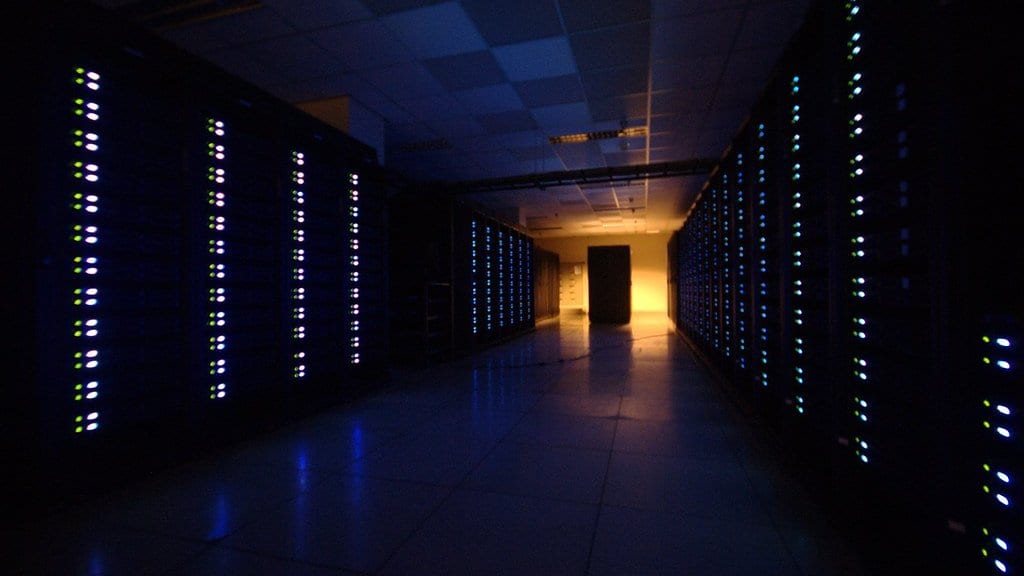 A dark data-centre aisle lined floor to ceiling with computer racks, their status lights glowing in rows on both sides