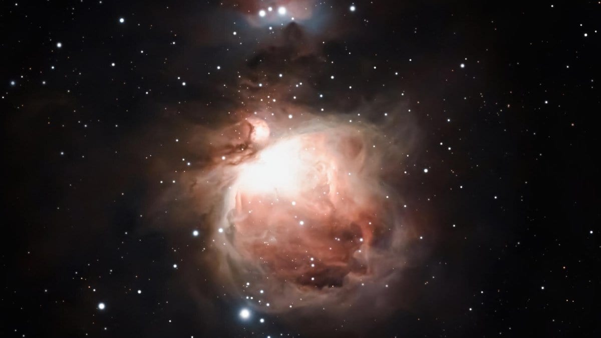 Deep telescopic photograph of the Orion Nebula: glowing pink and white clouds of gas and dust around a bright core, with scattered stars on a black sky.