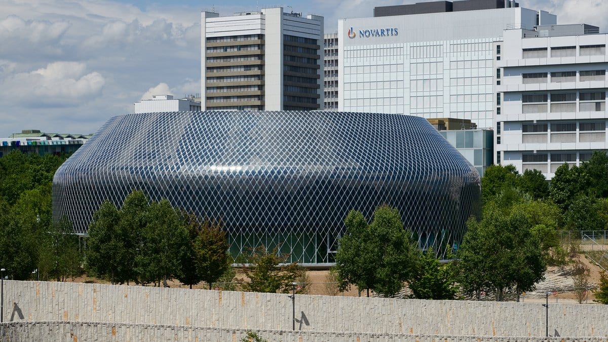 The silver, scale-clad Novartis Pavillon on the Novartis campus in Basel, Switzerland, with an office block carrying the Novartis name behind it.