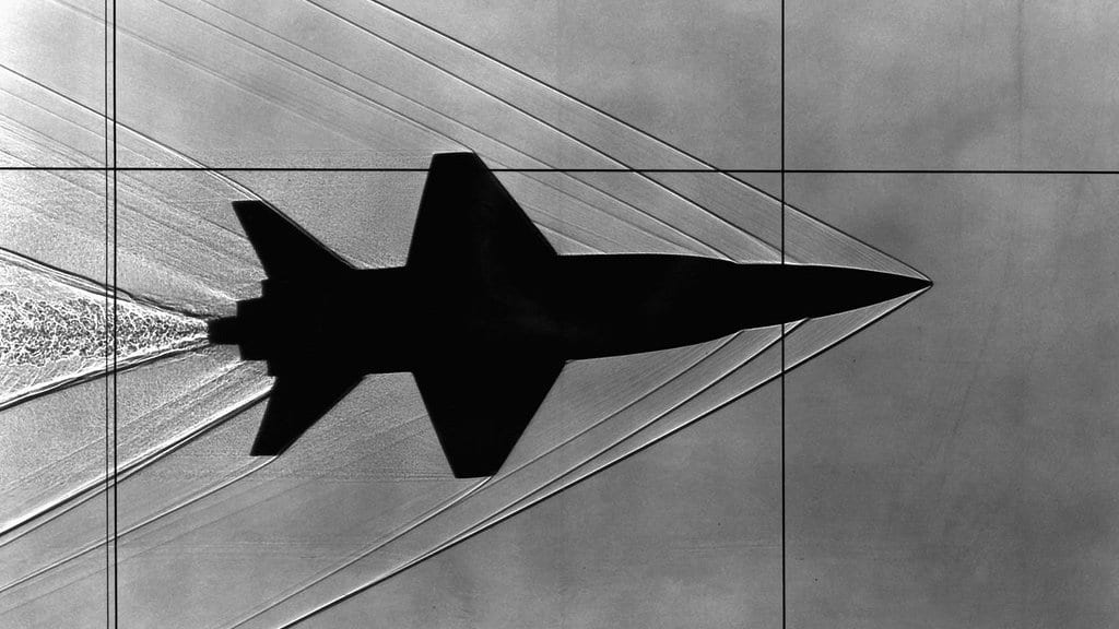 Black-and-white schlieren photograph of a wind-tunnel test: a dark aircraft model silhouette with sharp shock waves fanning out from its nose and tail and a mottled turbulent wake trailing behind it.