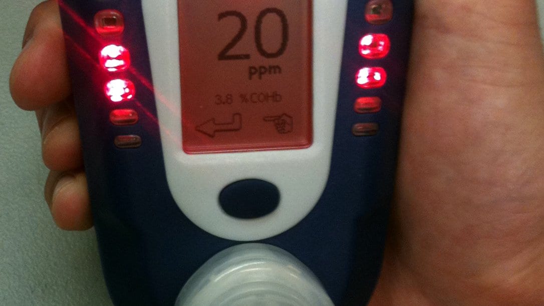 A hand holds a small breath carbon monoxide monitor whose screen reads 20 ppm and 3.8 percent COHb, with red indicator lights lit along both sides of the display.