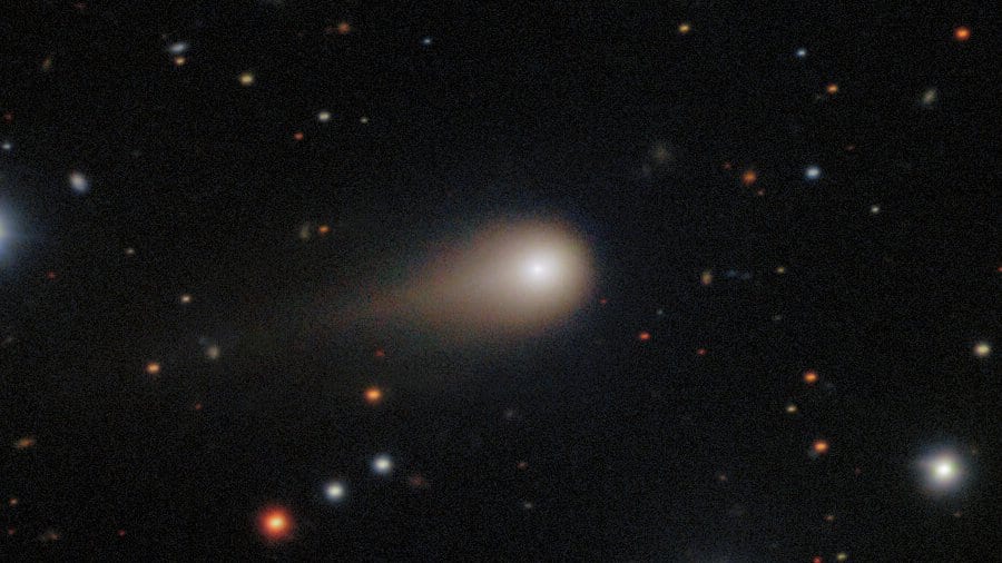 Colour telescope image of interstellar comet 3I/ATLAS: a bright condensed coma with a broad tail streaming to the left, set against a field of stars and small distant galaxies.