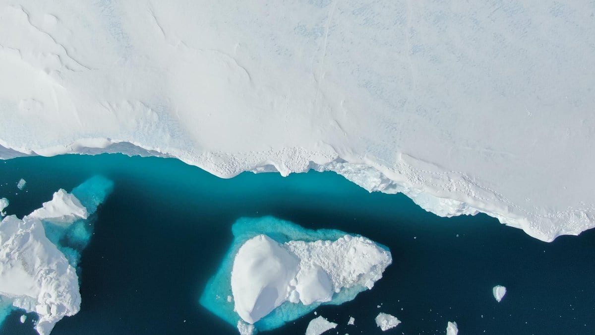 Straight-down aerial photograph of a white sea-ice sheet meeting dark blue open water, with a triangular ice floe and scattered smaller fragments drifting beyond the ice edge.