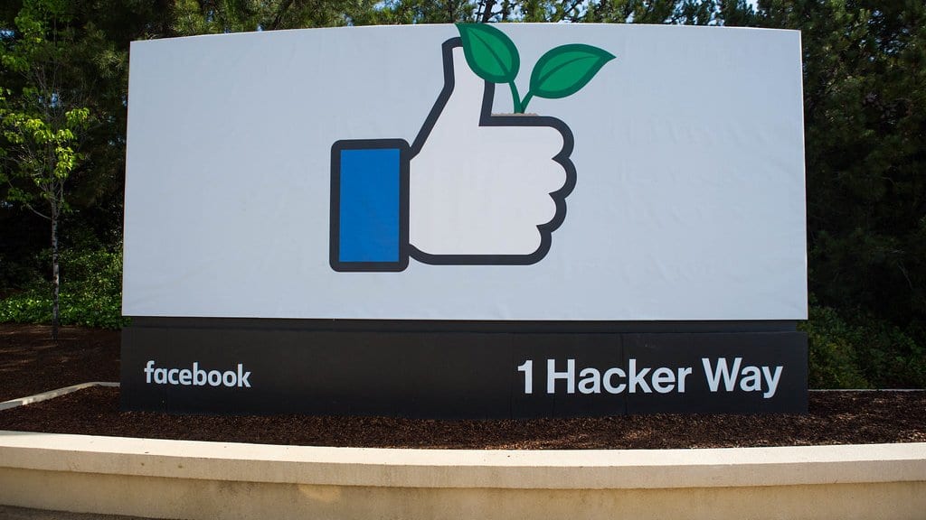 The thumbs-up entrance sign at 1 Hacker Way, Meta's headquarters campus in Menlo Park, California, with 'facebook' and '1 Hacker Way' lettering along its base.