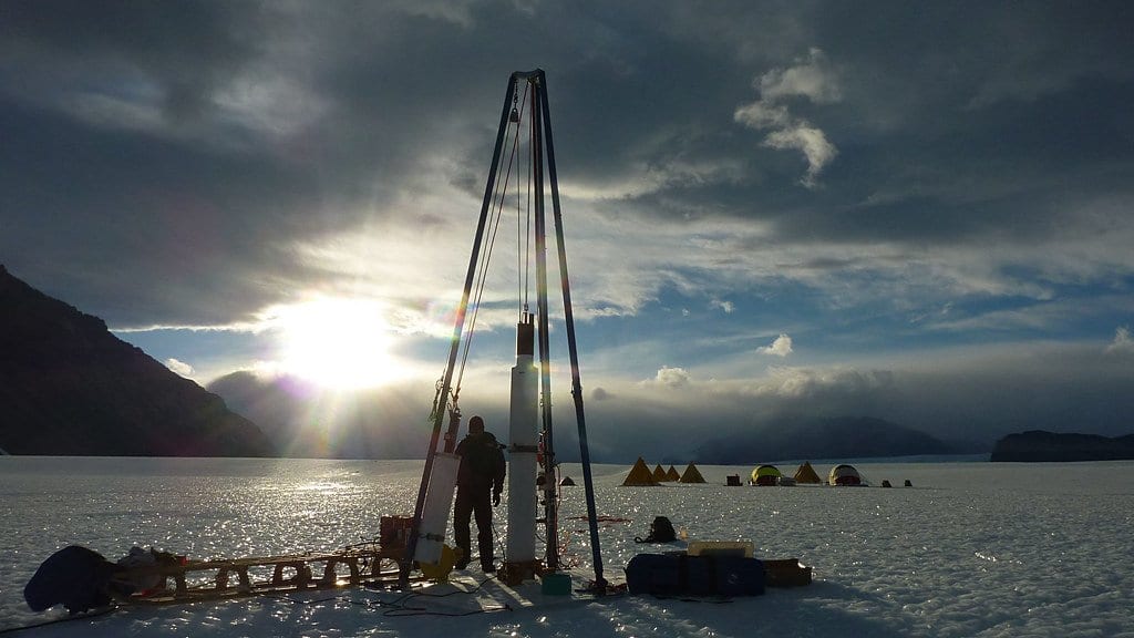 A tripod ice-core drill and a small field camp silhouetted on a snow-covered glacier surface, with the low sun behind them.