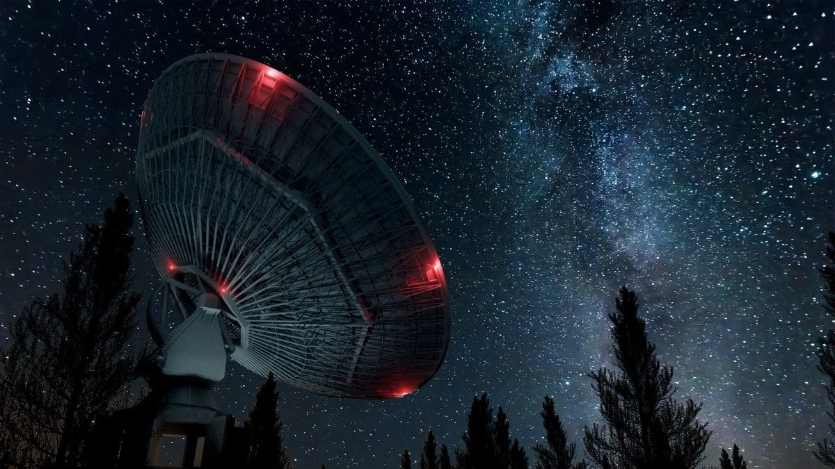 A large parabolic radio telescope dish silhouetted against a star-filled night sky, with the Milky Way rising behind it and dark trees along the horizon.