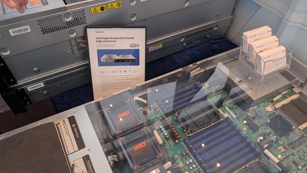 An opened rack-mount server in a glass display case at a trade show, its motherboard, memory slots and cooling fans visible, with a printed vendor spec card beside it.