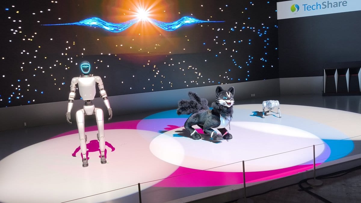 A silver bipedal humanoid robot standing on a white exhibition stage beside a robot cat and a small four-legged robot.