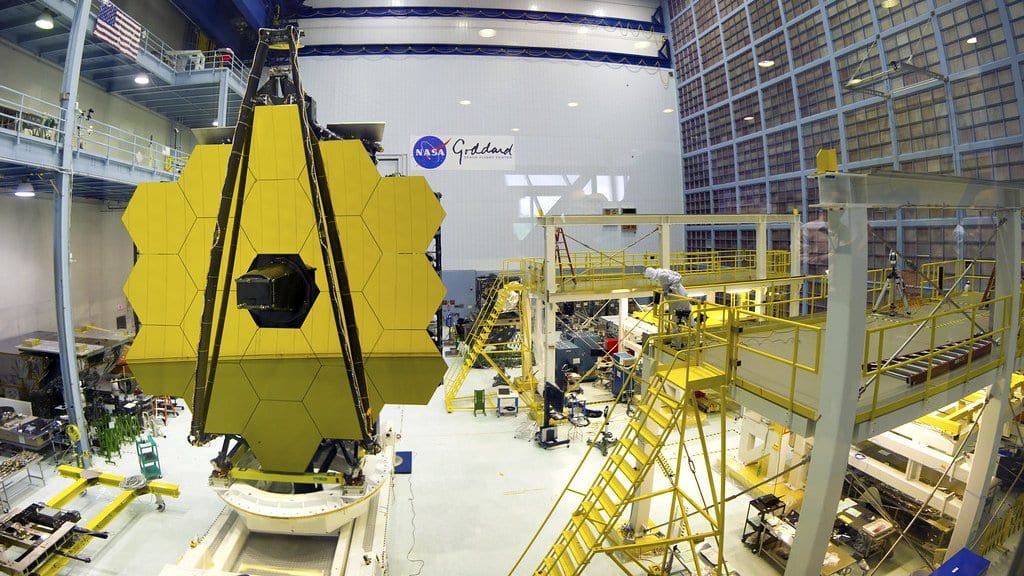 The gold-coated segmented primary mirror of the James Webb Space Telescope standing upright in a large clean room at NASA Goddard, with work platforms and engineers alongside.